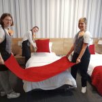 Trzy uczennice II klasy BS1S w zawodzie pracownik pomocniczy obsługi hotelowej z uśmiechami na twarzy, w strojach służbowych z przypiętymi kotylionami z bibuły w kształcie kwiatów doskonali swoje umiejętności praktyczne w zakresie ścielenia łóżek w pokoju hotelowym typu twin.
