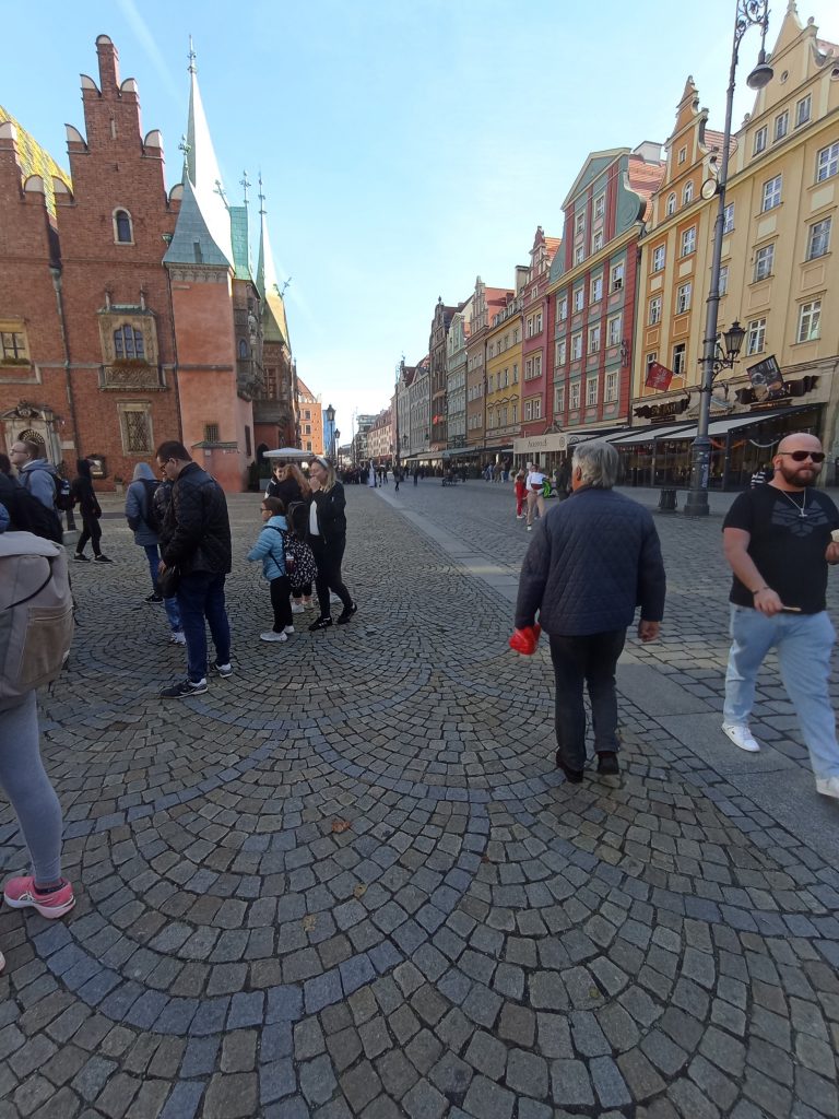 Grupa uczniów zwiedzających Rynek Wrocławski. W tle kamieniczki oraz inni przechodnie. Jest ładna słoneczna pogoda, czyste niebo.