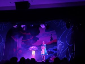 Alicja na scenie teatralnej, stoi obok purpurowego muchomora . Dekoracja w tle w kolorach niebiesko rożowych . Przyciemnione światła.  Alice on stage standing beside a purple mushroom. Alice is hugging herself close and looks intimidated.