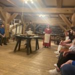 Gingerbread Museum. The musem chamber inside. In the middle there is a big wooden table with clay pots. Behind the table there are two people dressed in traditional folk costumes describing the procedure of making ginger bread. On both sides there are volunteers sitting on the wooden banches and listening to the presentation 