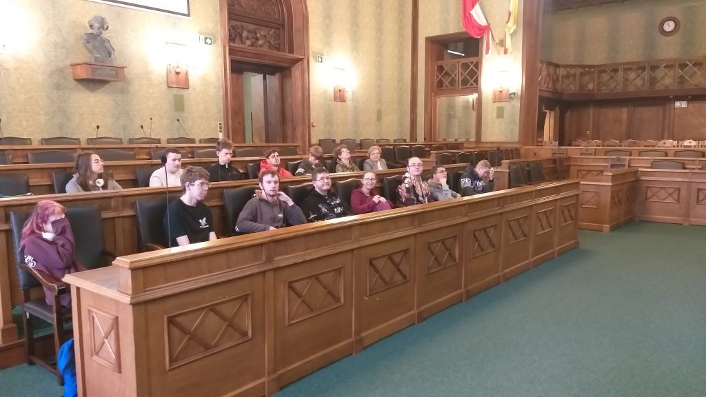 Wnętrze Sali sesyjnej Rady Miejskiej Wrocławia, na ścianie rozświetlone kinkiety. W lewym górnym rogu popiersie Ignacego Paderewskiego. W lożach (ławach, w których zasiadają radni miejscy) z pięknego brązowego drewna uczestnicy spotkania - uczniowie technikum ekonomicznego zwróceni twarzami do przemawiającego Pana Marcina Frąckiewicza – na zdjęciu niewidoczny. 