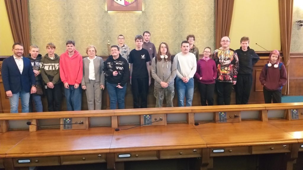 Na tle zabytkowego wnętrza Sali sesyjnej Rady Miejskiej Wrocławia grupa uczniów wraz z opiekunem i Gospodarzem spotkania zastępcą Dyrektora Biura Urzędu Miejskiego Panem Marcinem Frąckiewiczem – pierwszy od lewej i opiekunem wycieczki Panią Aleksandrą Grobelną – piąta od lewej. Uczniowie ubrani są w bluzy różnego koloru, szare, czerwone i białe. Na pierwszym planie fragment drewnianej brązowej ławy, w której podczas sesji zasiadają radni Wrocławia.