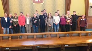 Na tle zabytkowego wnętrza Sali sesyjnej Rady Miejskiej Wrocławia grupa uczniów wraz z opiekunem i Gospodarzem spotkania zastępcą Dyrektora Biura Urzędu Miejskiego Panem Marcinem Frąckiewiczem – pierwszy od lewej i opiekunem wycieczki Panią Aleksandrą Grobelną – piąta od lewej. Uczniowie ubrani są w bluzy różnego koloru, szare, czerwone i białe. Na pierwszym planie fragment drewnianej brązowej ławy, w której podczas sesji zasiadają radni Wrocławia.