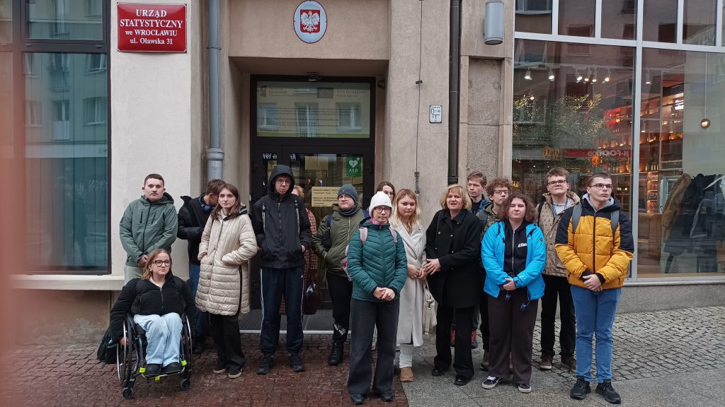 Od lewej w górnym rogu zdjęcia czerwona tablica z napisem Urząd Statystyczny we Wrocławiu ul Oławska 31. Na tle wejścia do budynku stoi grupa uczniów. Są ciepło ubrani w kurtkach różnego koloru. Jedna uczennica siedzi na wózku, pozostali pozują do zdjęcia stojąc. Wśród uczniów w czarnym okryciu opiekun Pani Ewa Kwiecień. Za plecami uczniów wejście do budynku z umieszczonym godłem Polskiej Rzeczpospolitej nad drzwiami. Po prawej stronie za plecami uczniów duże szklane okno.