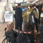 Holl Uniwersystetu Merito WSB we Wrocławiu. Na pierwszym planie grupa uczniów. Mają ciemne bluzy, jeden z nich (w jaśniejszej bluzie koloru khaki) na prawym ramieniu trzyma czarny plecak z dużym białym napisem Adidas. Grupę zamyka dwóch uczniów na wózkach. Zdjęcie ujmuje uczniów z tyłu. Są zgromadzeni przy stoisku informacyjnym, które jest po prawej stronie schodów prowadzących na I pietro. Przy stoisku duża tablica informacyjna z napisem UNIWERSYTET WSB MERITO WROCŁAW i dopisek mniejszymi literami wcześniej Wyższa Szkoła Bankowa. Holl jest jasno oświetlony.