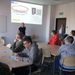 Uczniowie siedzą przy zaaranżowanych stolikach tworząc grupy. Mają różnokolorowe bluzy, przed sobą kartki oraz długopisy w ręku. Prowadząca warsztaty wykładowca akademicki Pani Dorota Otręba stoi przy tablicy multimedialnej twarzą do uczniów. Na slajdzie temat: „Deal or no Deal” zakreślony czerwoną obwódka i „Negocjacje, które działają nawet w szkole”. Na slajdzie jest również zdjęcie prowadzącej, logo uczelni i kod QR. W otwartych drzwiach, oparta o futrynę stoi Pani Aleksandra Grobelna - nauczyciel w ZSiP FECDiE we Wrocławiu.