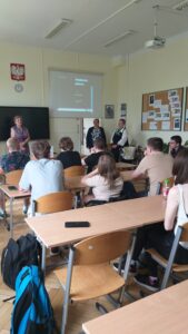 Uczniowie siedzą twarzami do prelegentów. Sa ubrani w różnokolorowe koszulki. Od lewej przed uczniami Pani Aleksandra Grobelna. Za Jej plecami na ścianie wygaszona tablica multimedialna, a nad tablicą godło Polski. Po prawej stronie twarzą do uczniów stoją nasi Goście, Panie Halina Chrzanowska i Renata Puch reprezentujące Zakład Ubezpieczeń Społecznych we Wrocławiu. Ponad głowami Pań
z ZUS-u widać rozwinięty ekran, na którym wyświetlana jest prezentacja o ZUS. Na ścianie po prawej stronie tablica korkowa z materiałami poglądowymi.